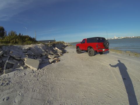 1995 Ford Bronco - A lifted red Ford Bronco is parked on a sandy beach next to the ocean. The vehicle has a black soft top and a spare tire mounted on the rea…