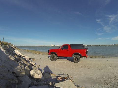 1995 Ford Bronco - A bright red Ford Bronco is parked on a sandy beach next to a body of water. The vehicle is viewed from the side, with a clear blue sky ove…