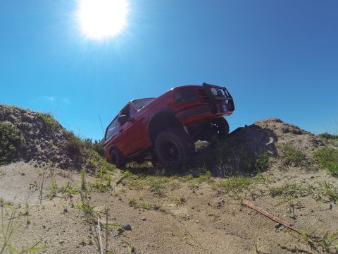 1995 Ford Bronco - A red 1995 Ford Bronco is shown from a low angle driving up a sandy hill under a bright blue sky. The vehicle has a brush guard on the fron…