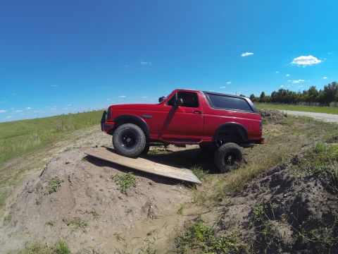 1995 Ford Bronco - A red Ford Bronco is parked on a dirt mound with a wooden plank under its front tire. The vehicle is facing left, with a clear blue sky and…