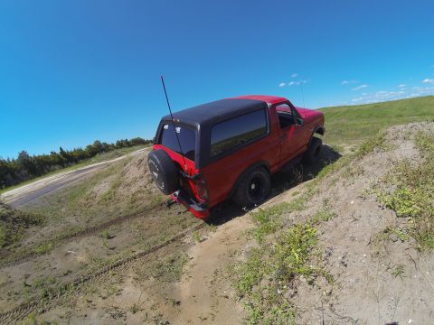 1995 Ford Bronco - A red 1995 Ford Bronco is shown from a rear-side angle, driving off-road on a dirt path with a spare tire mounted on the back. The vehicle…
