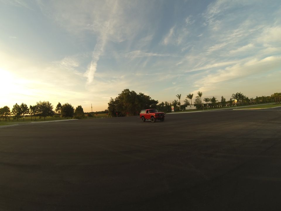 Red 1990s Ford Bronco parked on paved lot with trees and palm trees in background.
