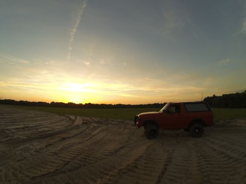 1995 Ford Bronco - A red 1995 Ford Bronco is parked on a dirt road with tire tracks, facing right, during a sunset. The sky is clear with a few wispy clouds a…