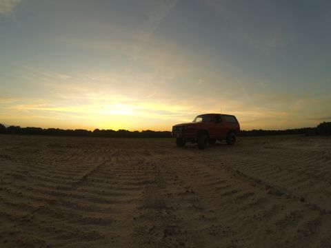 1995 Ford Bronco - A red Ford Bronco is parked in a dirt field during sunset. The sky is a gradient of yellow and blue with wispy clouds.