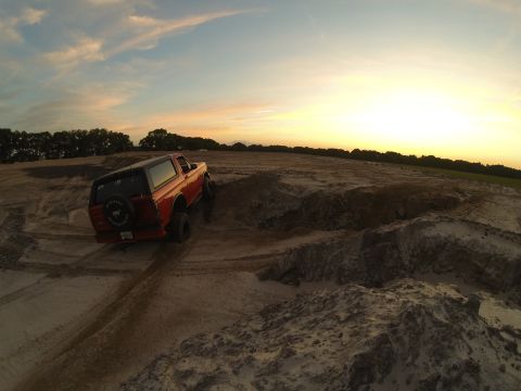 1995 Ford Bronco - A red 1995 Ford Bronco is shown from a rear, low angle, driving on a dirt track during sunset. The vehicle is in a construction or off-road…