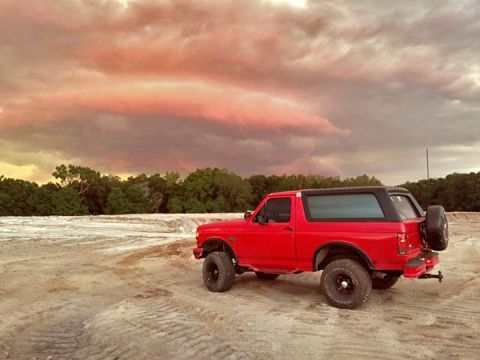 1995 Ford Bronco - A red 1995 Ford Bronco is parked in a dirt lot with a dramatic, colorful sky overhead.