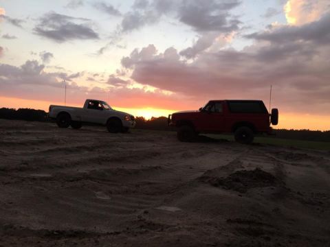 1995 Ford Bronco - Two off-road vehicles, a white pickup truck and a red SUV, are parked on a sandy terrain during sunset. The sky is filled with dramatic clo…