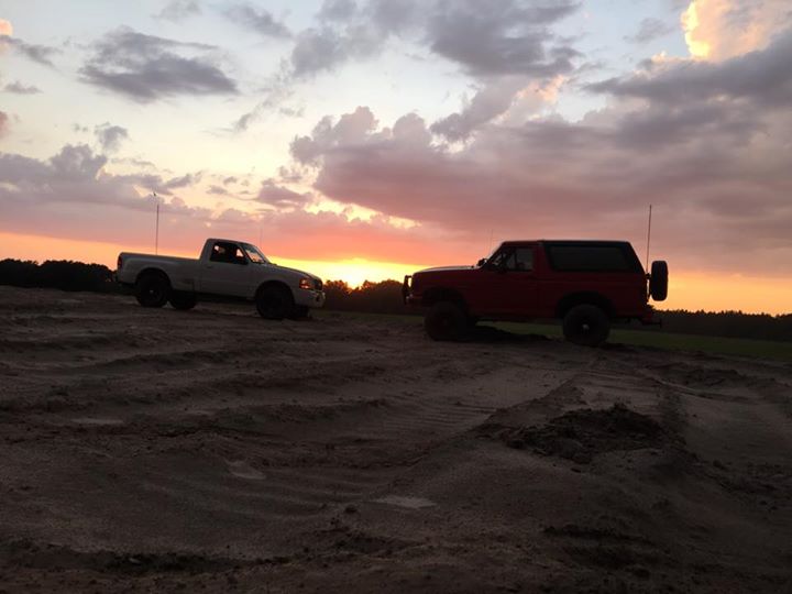 White pickup truck and red SUV parked on sand dunes at sunset.