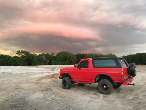 1995 Ford Bronco - A red 1995 Ford Bronco is parked on a dirt lot with a dramatic sky overhead. The SUV has a black roof and a spare tire mounted on the rear.