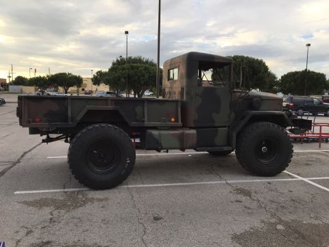 1989 Ford Bronco - A camouflage military-style truck with large off-road tires is parked in a parking lot under a cloudy sky.
