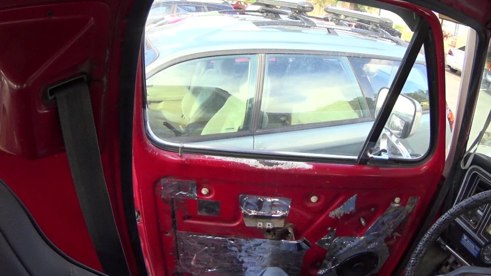 Interior of a red vehicle door with sound deadening material, showing a glimpse of another car through the window.