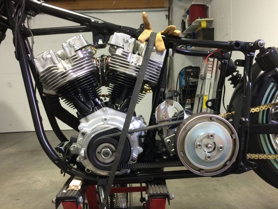 Custom motorcycle V-twin engine with belt drive on a stand, black frame, gold chain.