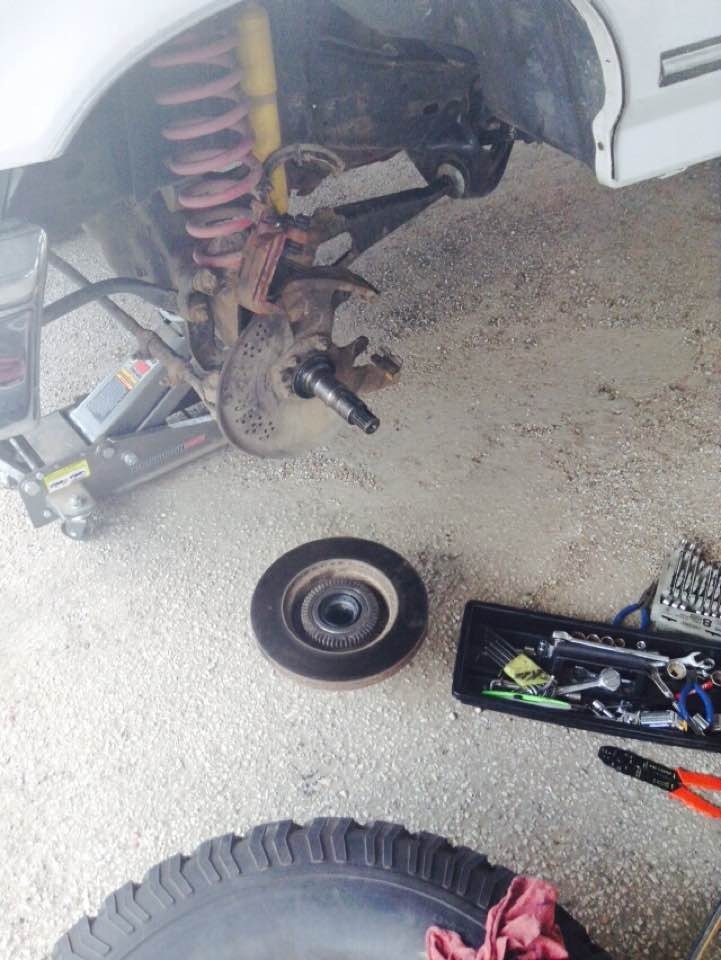 Disassembled vehicle front suspension with brake rotor, hub, toolbox, and tire on gravel.