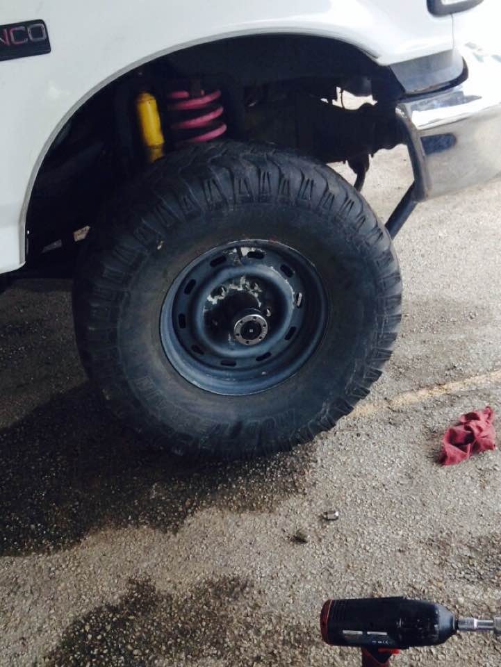 1996 Ford Bronco front wheel, suspension, yellow shock, pink spring, lug nuts, red rag on gravel.