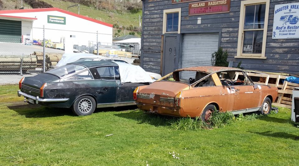 Two vintage coupes, one green and one rusty orange, parked on grass in front of a wooden building.