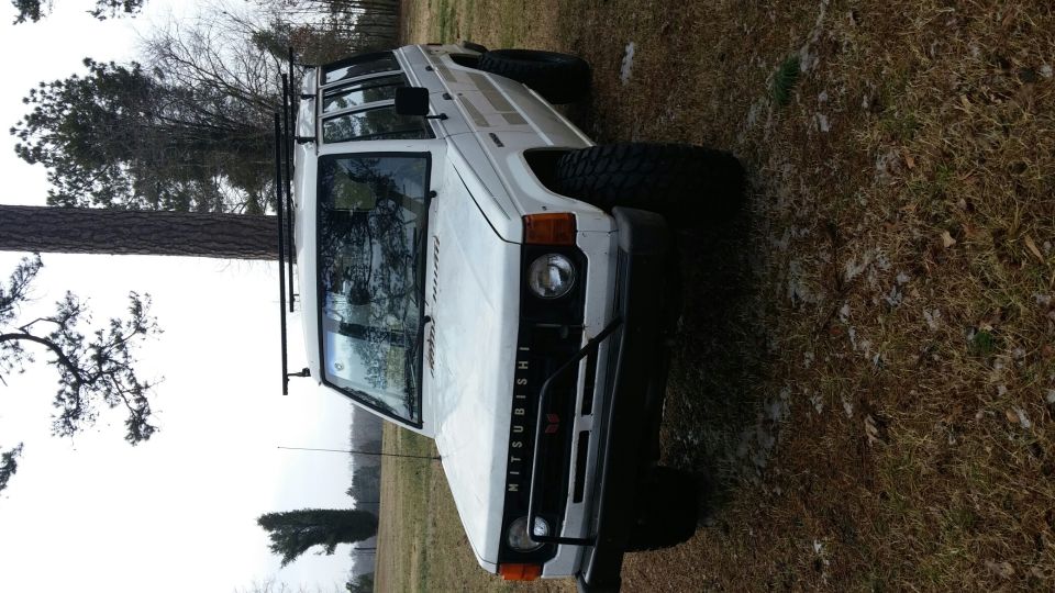 White Mitsubishi Montero SUV with roof rack and off-road tires parked outside.