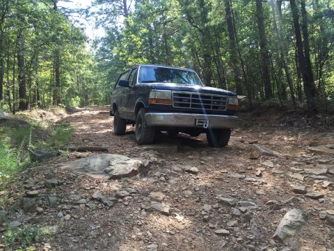 1993 Ford Bronco - A dark colored Ford Bronco is parked on a rocky, wooded trail. The front of the vehicle is visible, showing its grille, bumper, and tires.