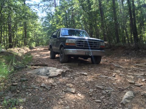 1993 Ford Bronco - A dark blue Ford Bronco is parked on a rocky, wooded trail. The front of the SUV is visible, showing its grille, headlights, and a winch on…