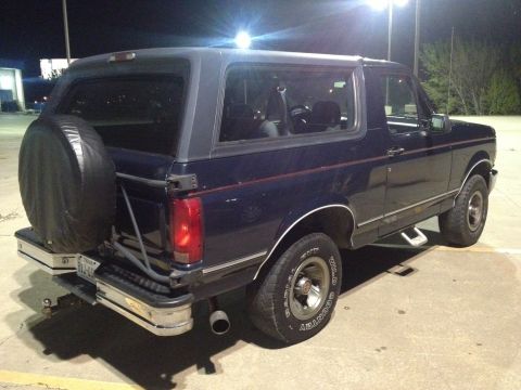 1993 Ford Bronco - The rear passenger side of a dark blue 1993 Ford Bronco is shown parked outdoors at night. A spare tire is mounted on the rear door, and a…