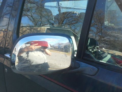 1993 Ford Bronco - A person is taking a photo of themselves in the side mirror of a vehicle, reflecting a street scene.