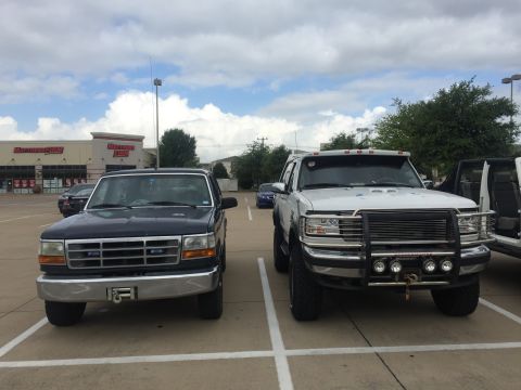 1993 Ford Bronco - Two Ford trucks are parked in a parking lot in front of a Mattress Firm store. The truck on the left is dark blue, and the truck on the rig…