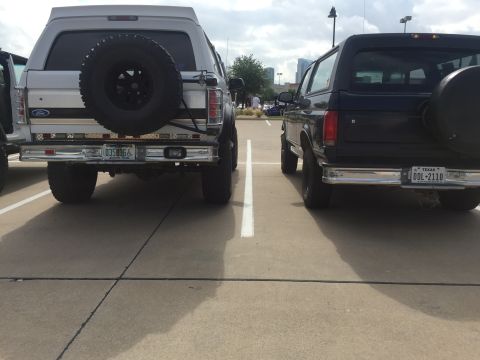 1993 Ford Bronco - The rear view of two Ford Broncos parked in a lot. The Bronco on the left is silver with a spare tire mounted on the back, and the Bronco o…