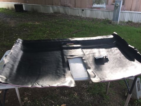 1993 Ford Bronco - A black headliner is laid out on a table outdoors, showing its textured surface and some creases. A small electronic device is placed on th…