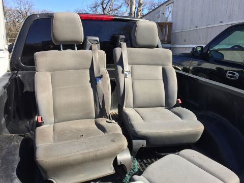 1993 Ford Bronco - Two front bucket seats with headrests and seatbelts are loaded in the bed of a pickup truck. A black pickup truck is parked next to the sea…