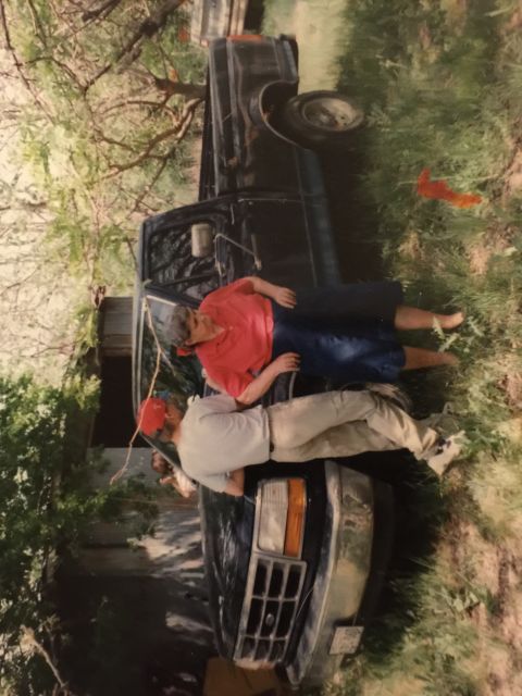 1993 Ford Bronco - Two people are interacting with the front of a dark-colored Ford Bronco, which is parked outdoors on a grassy area with trees in the backgr…
