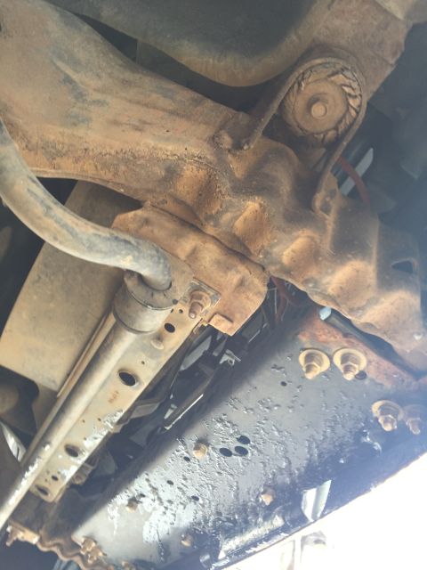 1993 Ford Bronco - Underside view of a rusty suspension component, possibly a sway bar link, attached to the frame of a vehicle. The frame appears to be paint…