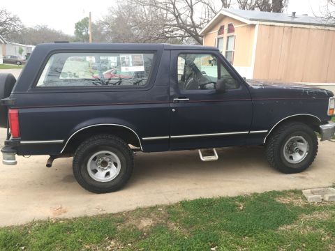 1993 Ford Bronco - The side profile of a dark blue Ford Bronco is shown parked on a concrete driveway with grass and trees in the background. The vehicle has…