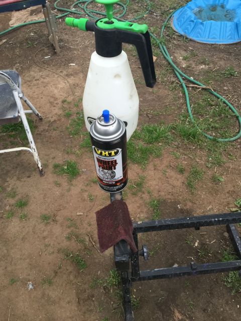1993 Ford Bronco - A can of VHT wheel paint sits next to a garden sprayer and a metal stand with a maroon pad on it, set against a dirt and grass background.