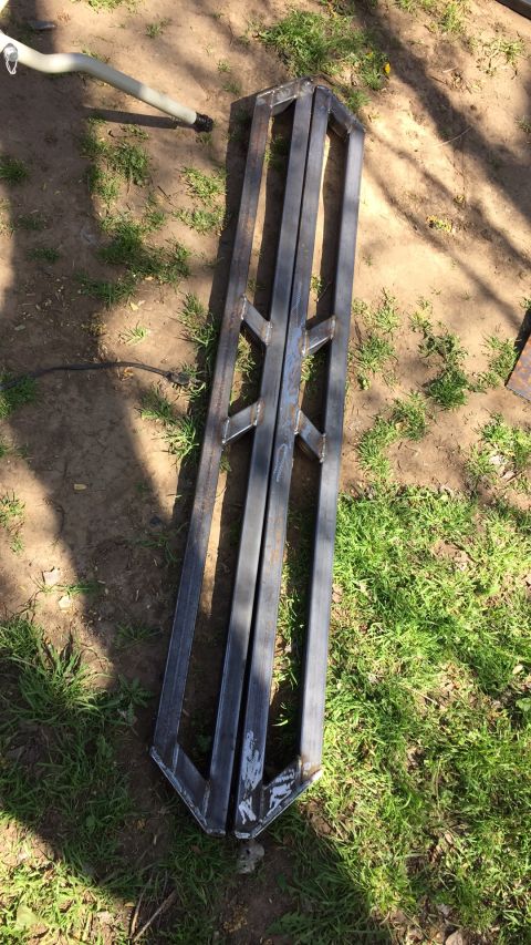 1993 Ford Bronco - Two custom-fabricated metal frames are laid out on the ground, partially covered by grass and dirt. A light-colored pipe with a black fitti…