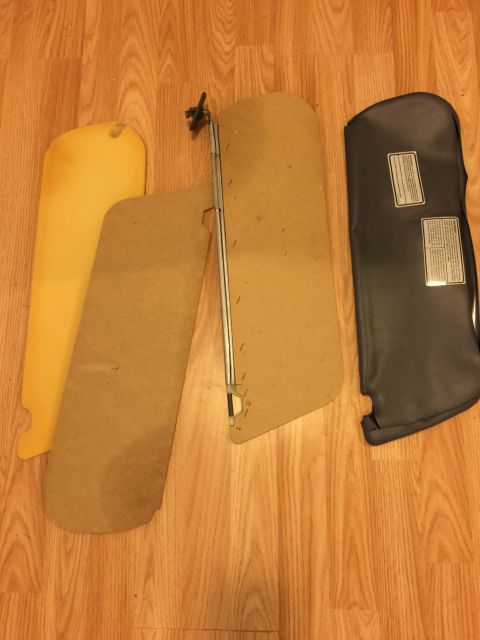 1993 Ford Bronco - Three sun visors are laid out on a wooden floor. Two are tan cardboard with metal supports, and one is a dark gray vinyl-covered visor with…