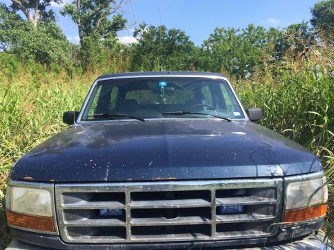 1993 Ford Bronco - The front view of a dark blue Ford Bronco is parked in tall green grass with trees in the background. The vehicle has a chrome grille and o…
