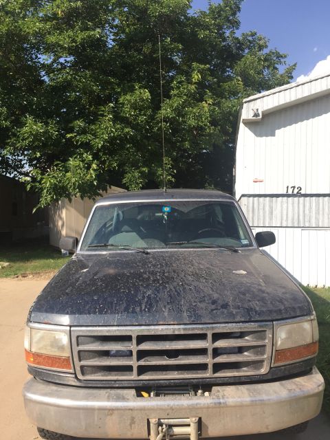 1993 Ford Bronco - Front view of a dusty, dark blue Ford truck with a chrome bumper and a winch. The truck is parked in front of a white building with the num…