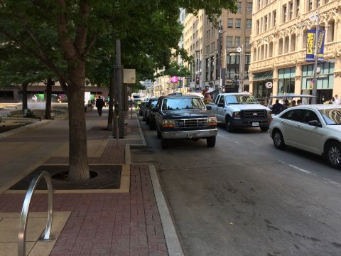 1993 Ford Bronco - A line of vehicles, including a black Ford pickup truck, is parked on a city street next to a sidewalk with trees. A white sedan is visible…