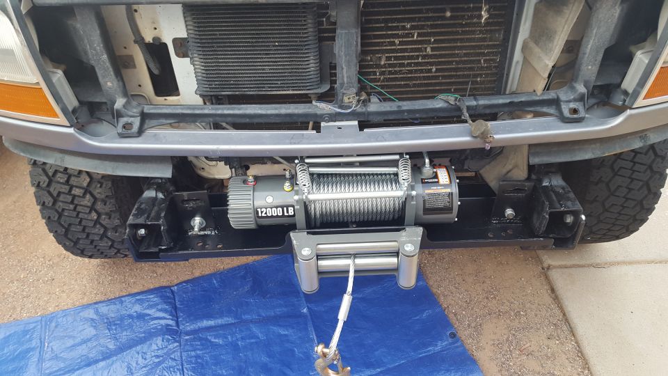 12000 LB winch mounted on front of white Ford with bumper removed, cable hanging onto blue tarp