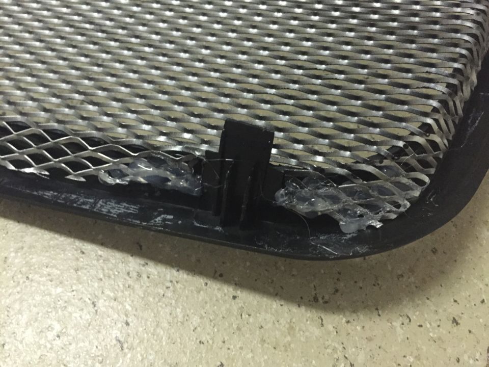 Black plastic grille frame with metal mesh insert attached with clear adhesive and clip.