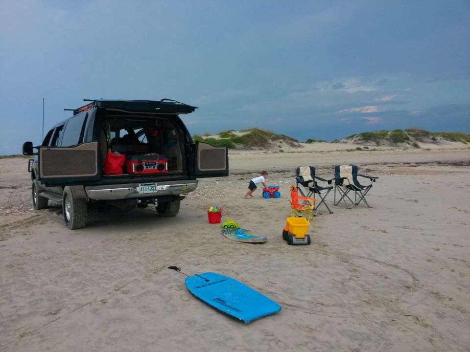 Black Ford Excursion parked on a beach with open hatch, beach chairs, and toys.