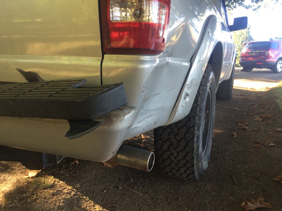Rear passenger side of a light-colored pickup truck with a damaged bumper and off-road tire. Red SUV in background.