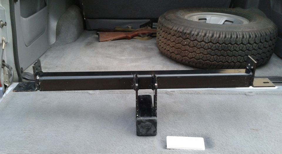 Black trailer hitch assembly lying in gray-carpeted SUV cargo area next to a spare tire
