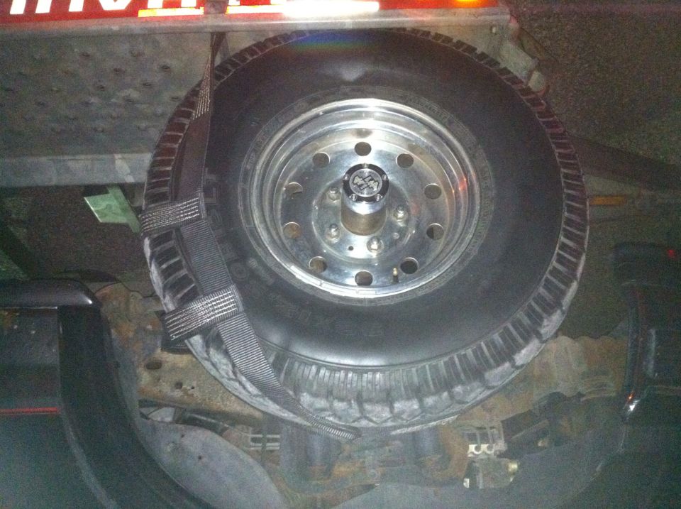 Spare tire with chrome rim and 4x4 center cap secured by straps to a trailer.