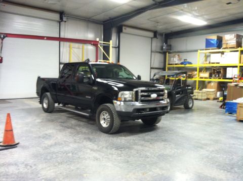 2004 Ford F-250 - A black Ford F-250 pickup truck is parked inside a garage next to a utility vehicle. The truck is viewed from a 3/4 front angle, showing it…