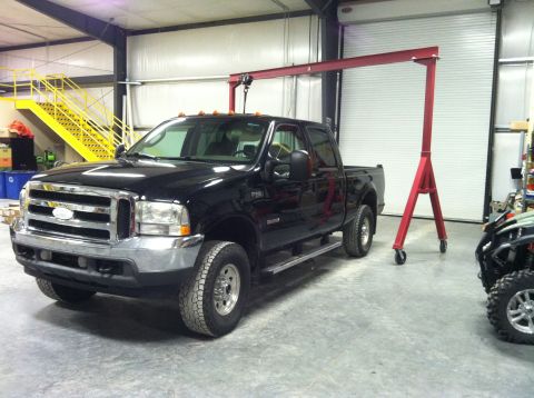 2004 Ford F-250 - A black Ford F-250 pickup truck is parked inside a garage or workshop, with a red gantry crane positioned over it. A yellow staircase is vi…