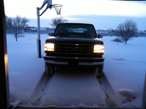2004 Ford F-250 - A black Ford pickup truck with its headlights on sits in a snowy driveway, facing the camera. A basketball hoop is visible to the left of t…