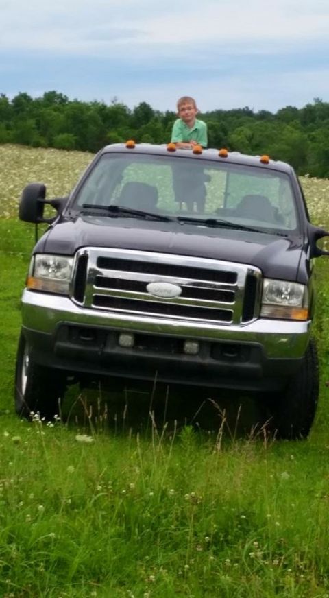 2004 Ford F-250 - A young boy stands on the roof of a dark-colored Ford F-250 pickup truck parked in a grassy field with trees in the background.