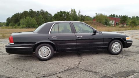 2001 Ford Crown Victoria - Side view of a dark-colored sedan parked outdoors on a cracked paved surface.
