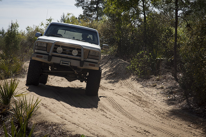 White 1995 Ford Bronco driving on a sandy trail with brush and trees