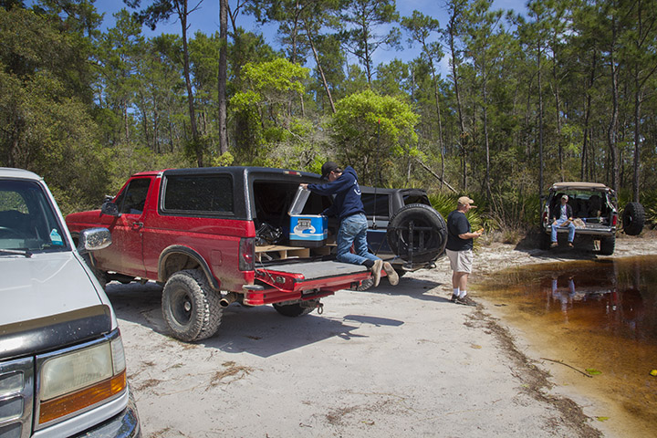 Red 1995 Ford Bronco with its tailgate down, someone loading a cooler. Another Bronco and a white truck nearby